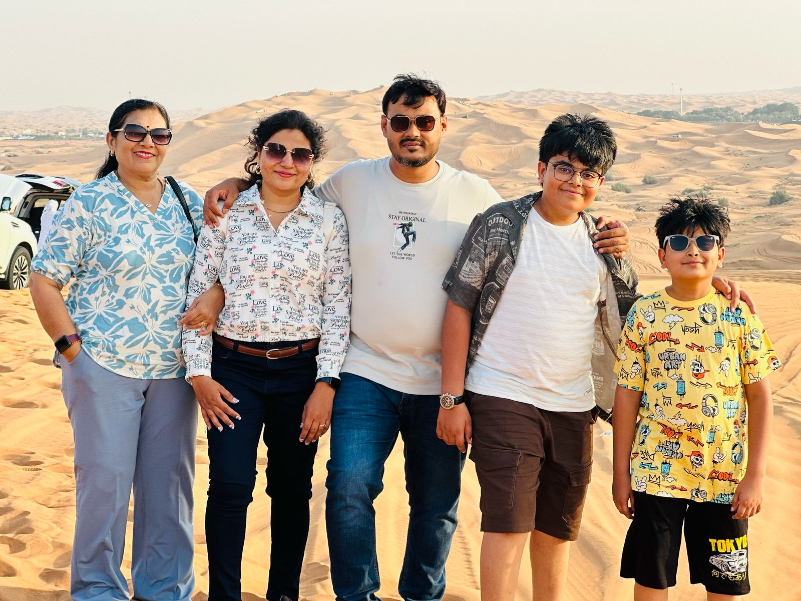 A family of five posing together on sandy desert dunes, smiling at the camera with a parked SUV and rolling dunes in the background under a clear sky.